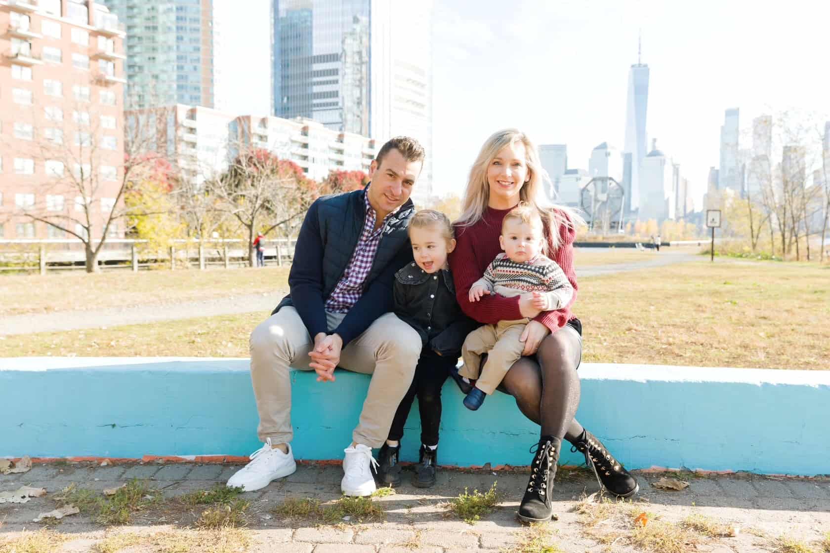 Family sitting on a park ledge with city skyscrapers and skyline in the background.